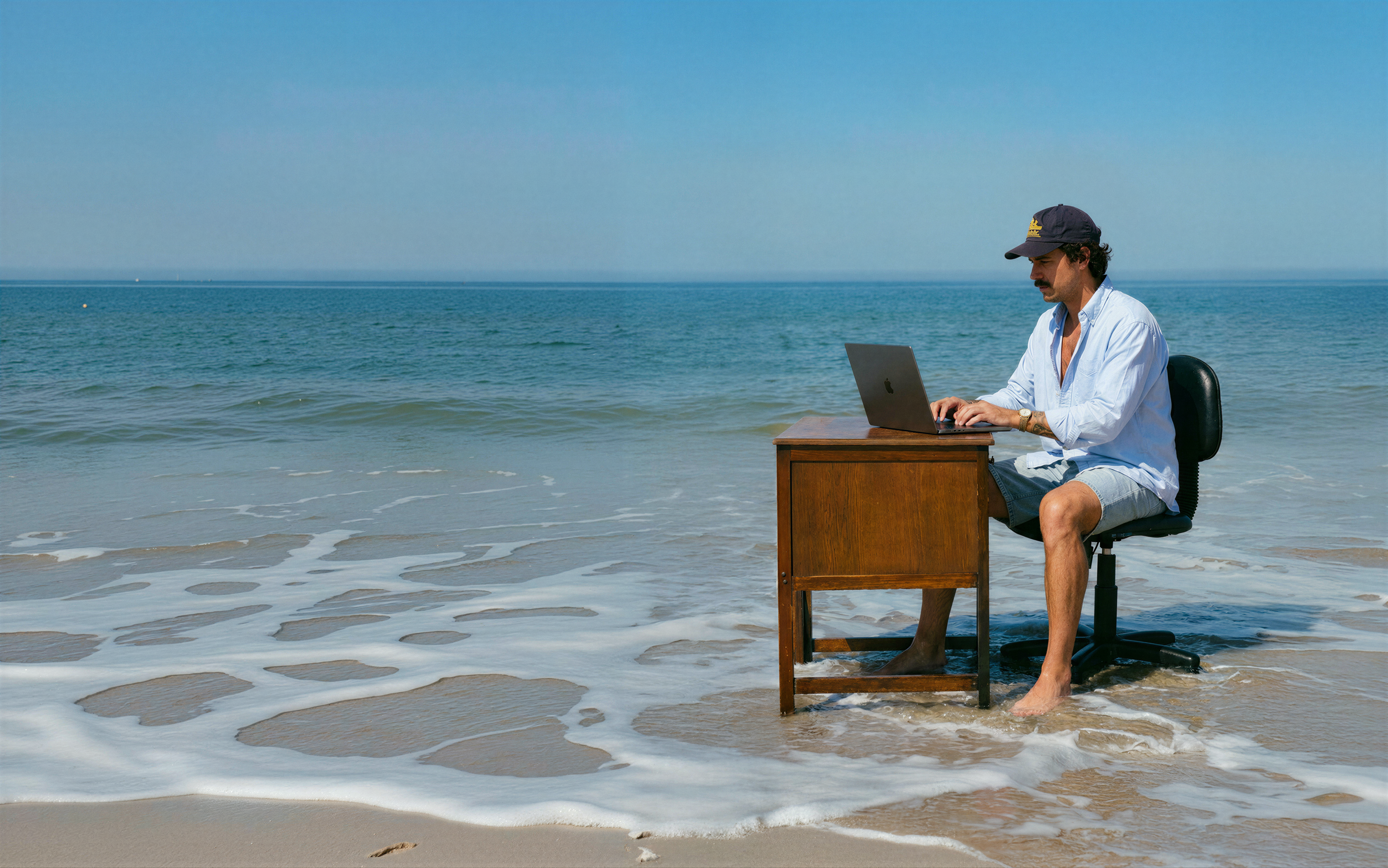 A man working on a laptop at a wooden desk that stands in the sea, at the edge of a beach