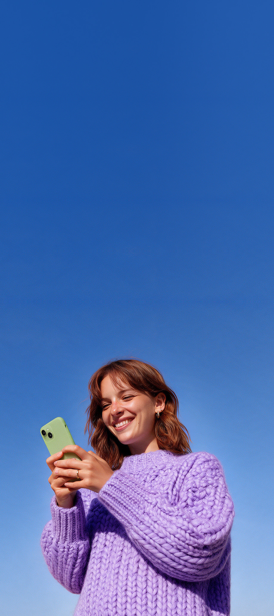 A woman in a lilac jumper under a clear blue sky, smiling at her phone